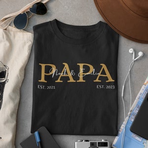 Puede incluir: Camiseta negra con letras doradas que dicen "PAPA Noah & Emilia EST. 2021 EST. 2023".