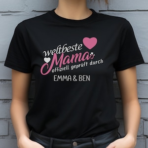 Könnte beinhalten: Schwarzes T-Shirt mit pinkfarbenem Text, der "weltbeste Mama" mit einem pinkfarbenen Herz und einem weißen Herz lautet. Der Text setzt sich mit "offiziell geprüft durch" und "EMMA & BEN" in weiß fort.