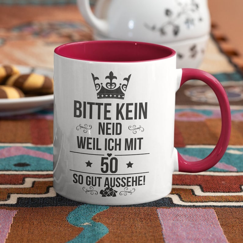 Pode incluir: Caneca de cer&acirc;mica branca com interior e al&ccedil;a bord&ocirc;. A caneca apresenta um texto preto que diz "BITTE KEIN NEID WEIL ICH MIT 50 SO GUT AUSSEHE!" com uma coroa e detalhes florais. A caneca est&aacute; sobre uma superf&iacute;cie estampada.