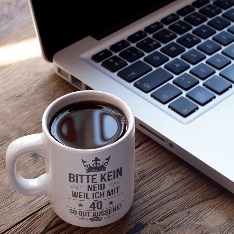 May include: White ceramic mug with black text that reads "Bitte kein Neid weil ich mit 40 so gut aussehe!"  The mug is filled with coffee and sitting on a wooden table next to a silver laptop.