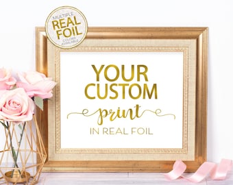 May include: A gold framed picture with the text "YOUR CUSTOM print IN REAL FOIL" in gold foil lettering. The frame is sitting on a white wooden surface with pink ribbon and a gold vase with pink flowers.