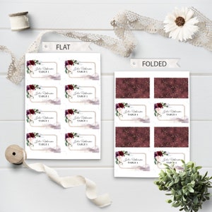 May include: Printable place cards with a burgundy floral design and gold accents. The cards are shown flat and folded. The text on the cards reads "Julia Robinson, Table 1".