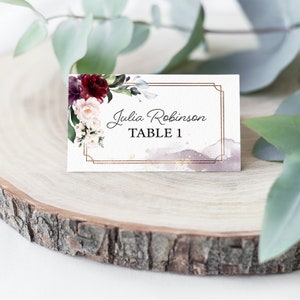May include: A white wedding place card with a gold frame and a watercolor background. The card features a floral design with burgundy, pink, and white flowers. The text on the card reads "Julia Robinson Table 1".