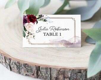 May include: A white wedding place card with a gold frame and a watercolor background. The card features a floral design with burgundy, pink, and white flowers. The text on the card reads "Julia Robinson Table 1".