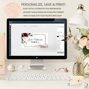 May include: A computer screen displaying a digital design of a table number card with a floral design and the text "Julia Robinson Table 1". The design is in shades of pink, burgundy, and gold. The screen is on a desk with a keyboard, a lamp, and other office supplies.