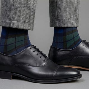 May include: A pair of black leather oxford shoes with cap toes and black laces, worn with blue, green, and black plaid socks and grey herringbone trousers. The shoes feature a smooth finish, a low heel, and a thin brown sole edge detail.