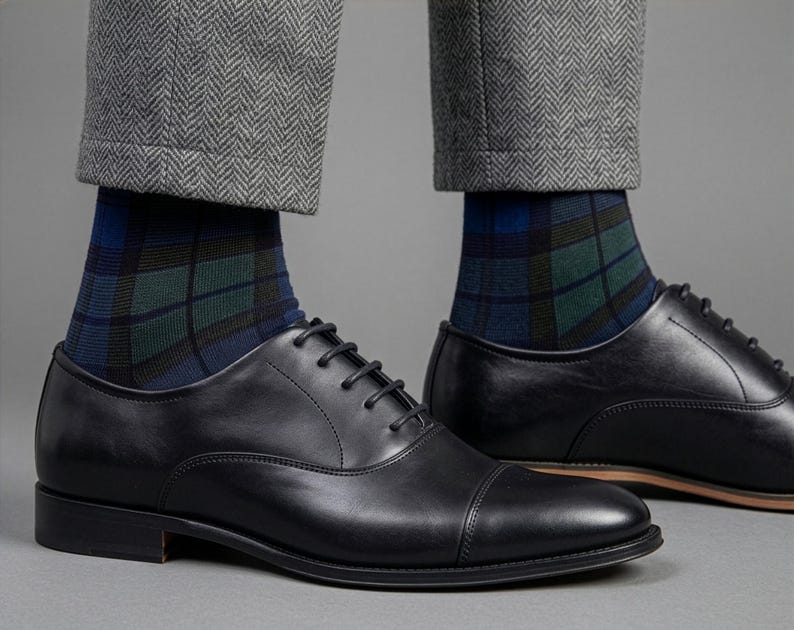 May include: A pair of black leather oxford shoes with cap toes and black laces, worn with blue, green, and black plaid socks and grey herringbone trousers. The shoes feature a smooth finish, a low heel, and a thin brown sole edge detail.