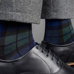 May include: A pair of socks featuring a blue, green, and black plaid pattern, worn with black leather oxford shoes and grey herringbone trousers. The socks extend above the ankle, showcasing a detailed grid design against the dark footwear.