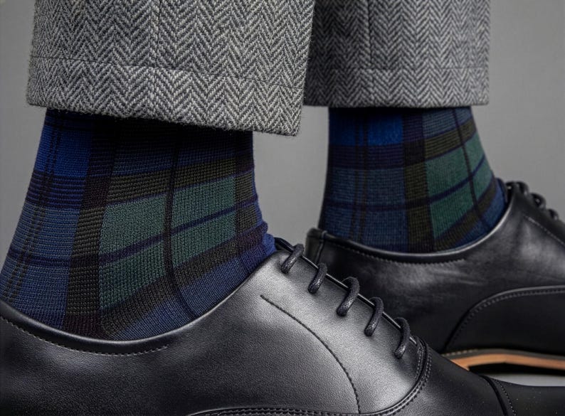 May include: A pair of socks featuring a blue, green, and black plaid pattern, worn with black leather oxford shoes and grey herringbone trousers. The socks extend above the ankle, showcasing a detailed grid design against the dark footwear.