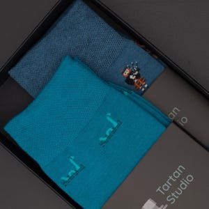Könnte beinhalten: Zwei Paar Socken in einer schwarzen Box. Ein Paar ist blau mit einem weißen und blauen Muster. Das andere Paar ist türkis mit einem weißen und türkisfarbenen Muster. Die Box hat ein Etikett mit der Aufschrift "Tartan Studio".