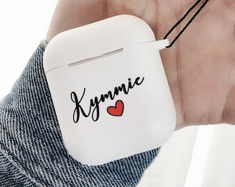 May include: White Airpods case with a black strap and the name "Kymmi" written in black cursive with a red heart.