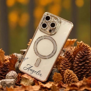 May include: A white phone case with a gold ring and a gold camera lens cover. The case has a sparkly finish and the name "Angelia" is written on the back.