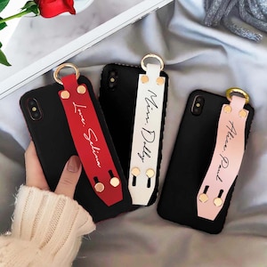 Puede incluir: Tres fundas de teléfono negras con correas personalizadas. Las correas son rojas, blancas y rosas con detalles dorados. Las correas tienen los nombres "Love: Selina", "Miss: Dolly" y "Alice: Paul" escritos en ellas.