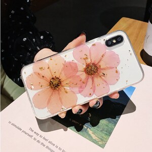May include: A clear phone case featuring a pressed flower design. The flowers are pink with gold accents. The case is designed for a smartphone with a dual camera lens.