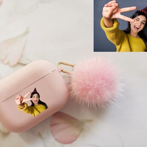 May include: A pink Airpods case with a fuzzy pink pom-pom keychain. The case has a photo of a person wearing a party hat and making a peace sign.
