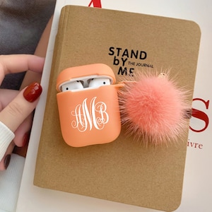 May include: A peach-coloured Airpods case with a white monogram "AMB" and a fluffy peach-coloured pom-pom keychain. The case is sitting on a brown notebook with the text "Stand By Me The Journal" printed on it.