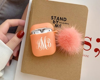 May include: A peach-colored Airpods case with a white monogram "AMB" and a fluffy peach-colored pom-pom keychain. The case is sitting on a brown notebook with the text "Stand By Me The Journal" printed on it.