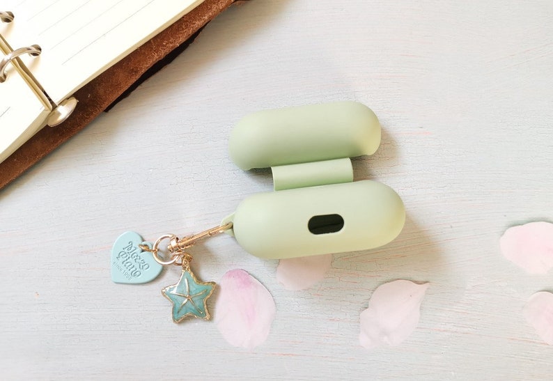 May include: A light green silicone case for wireless earbuds with a gold chain and charm attached. The charm is a blue starfish with a heart-shaped tag that says "Alice & Luna".