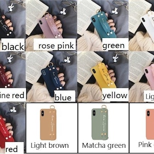 May include: Ten different colored phone cases with a strap and a small ring. The cases are made of a soft, textured material and have a handwritten-style inscription that says "Love Yourself". The colors are black, rose pink, green, white, wine red, blue, yellow, light pink, red, light brown, matcha green, and pink orange.