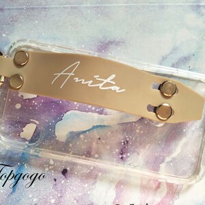 May include: A clear phone case with a beige phone grip attached. The grip has the name "Anita" written in white lettering. The case is on a background with a watercolour design.