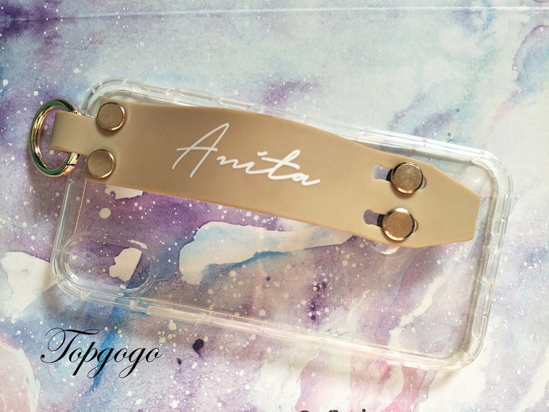 May include: A clear phone case with a beige phone grip attached. The grip has the name "Anita" written in white lettering. The case is on a background with a watercolour design.