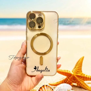 May include: A white iPhone case with a gold ring holder and a gold camera lens cover. The case has the name "Angelia" written on it in black cursive with two small hearts above the name.