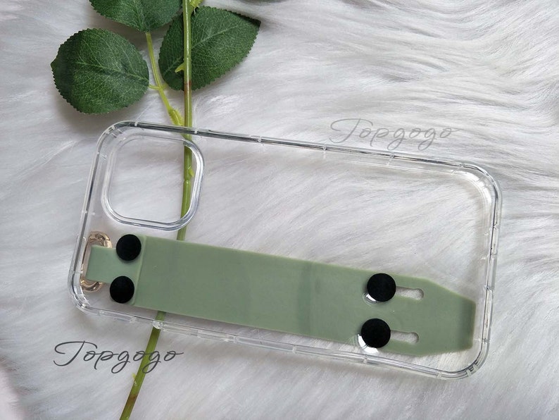 May include: Clear phone case with a sage green strap and black circular accents. The case has a gold-colored metal loop and the text "Topgogo" is visible. The case is designed to protect a phone.