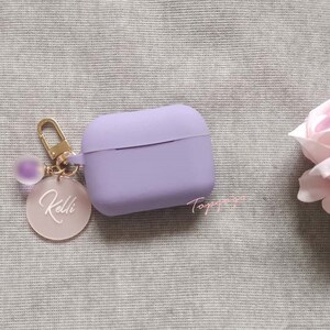 May include: A lavender silicone case for AirPods with a gold keychain featuring a clear charm with the name "Kelli" written in white.