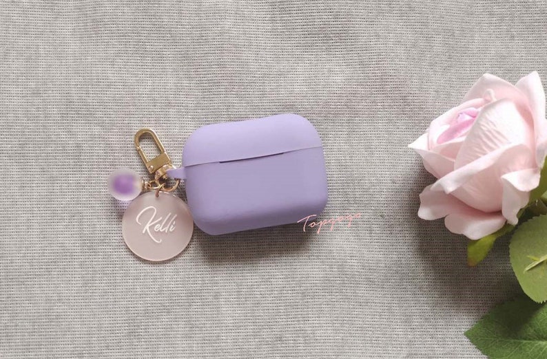 May include: A lavender silicone case for AirPods with a gold keychain featuring a clear charm with the name "Kelli" written in white.