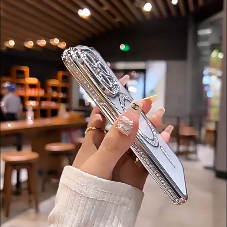 Op de afbeelding: Een witte telefoonhoes met een zilveren, met juwelen bezet ontwerp. De hoes heeft een verhoogd, versierd ontwerp met strass steentjes en een transparante achterkant.