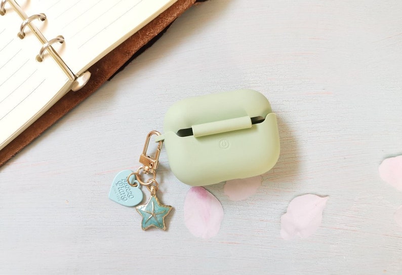 May include: A light green silicone case for wireless earbuds with a gold chain and charm. The charm is a blue starfish with a heart-shaped charm that says "Good Vibes".