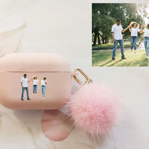 May include: A pink Airpods case with a fuzzy pink pom-pom keychain. The case has a photo of a family of three, a man and two girls, holding hands and smiling.
