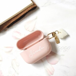 May include: A pink Airpods case with a white pearl heart charm attached to a gold chain. The case has the name "Sindu" written on it in white.
