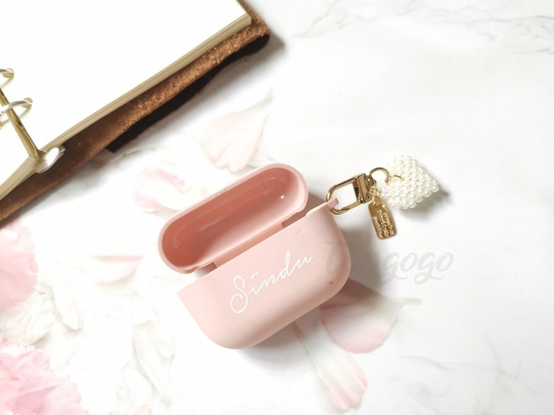 May include: A pink Airpods case with a white pearl heart charm attached to a gold chain. The case has the name "Sindu" written on it in white.