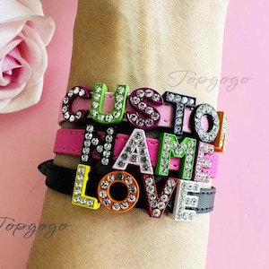May include: A stack of colorful bracelets spelling out "CUSTOM NAME LOVE" with rhinestone-studded letters. The bracelets are on pink and black leather bands. A pink rose is in the background.