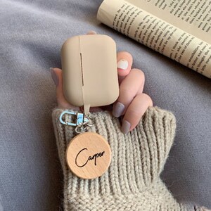 May include: A beige silicone case for wireless earbuds with a wooden keychain that says "Carper".