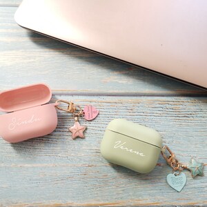 May include: Two personalized Airpod cases with charms. The pink case has the name "Sindu" and the green case has the name "Verena". Both cases have a gold chain with a star charm and a heart charm.