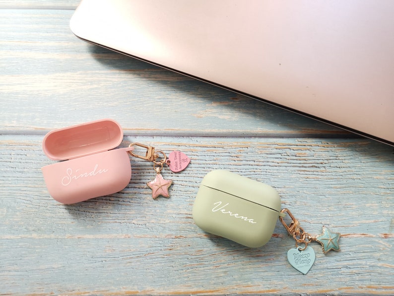 May include: Two personalized Airpod cases with charms. The pink case has the name "Sindu" and the green case has the name "Verena". Both cases have a gold chain with a star charm and a heart charm.