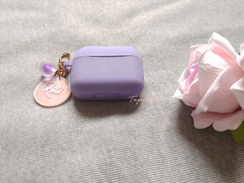 May include: A lavender silicone case for wireless earbuds with a gold chain and a pink charm with the word "Kelli" engraved on it.