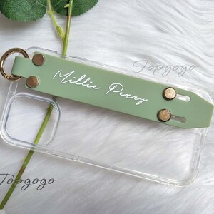 May include: A clear phone case with a sage green strap featuring the name "Millie Perry" in white script. The strap has gold-colored rivets and a gold ring. The case is shown on a white, fluffy surface.