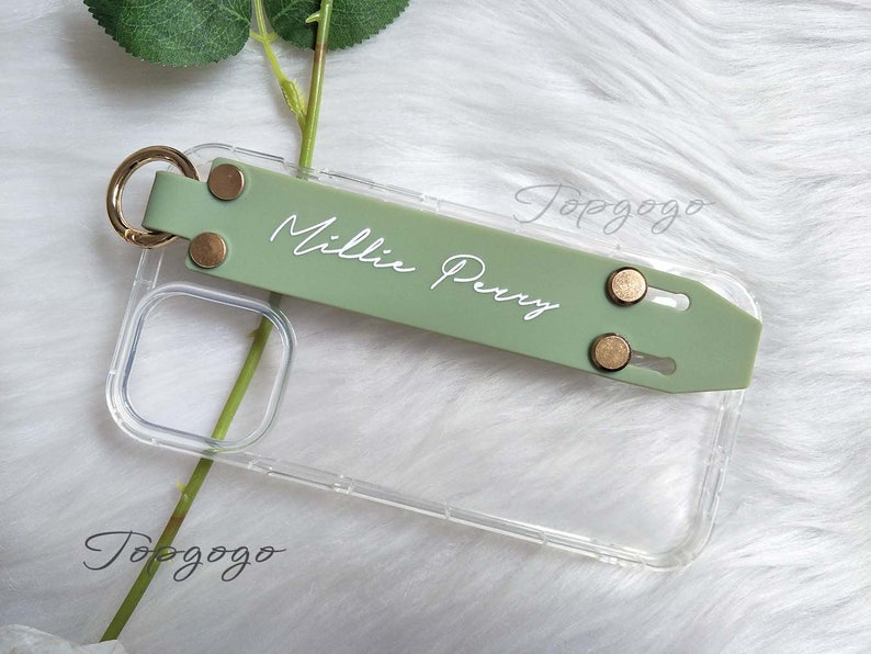 May include: A clear phone case with a sage green strap featuring the name "Millie Perry" in white script. The strap has gold-colored rivets and a gold ring. The case is shown on a white, fluffy surface.