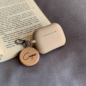 May include: A beige Airpod case with a silver keychain attached to a wooden keychain charm with the word "Carper" engraved on it.
