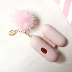May include: A pink fuzzy keychain charm attached to a pink silicone case for wireless earbuds. The case has a small opening for charging.