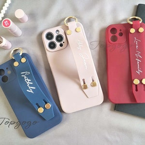 Peut inclure: Trois étuis de téléphone avec une sangle et un anneau doré. Les étuis sont rose, rouge et bleu. Les sangles portent les mots "Abby Jean", "Love My Family" et "Beautifully" écrits dessus.