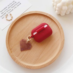 May include: Red silicone case for wireless earbuds with a gold heart-shaped charm attached to a gold chain. The case is sitting on a wooden surface.