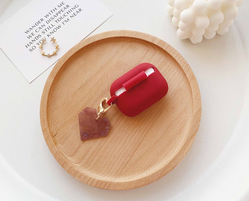 May include: Red silicone case for wireless earbuds with a gold heart-shaped charm attached to a gold chain. The case is sitting on a wooden surface.