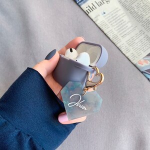 May include: A gray silicone case for wireless earbuds with a gold heart-shaped charm attached. The charm has a clear, iridescent heart-shaped tag with the word "Jhon" written on it.