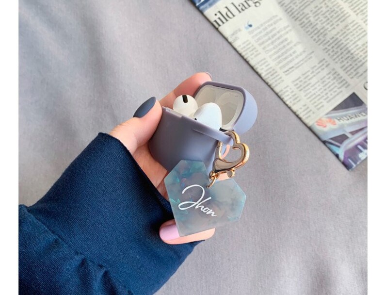 May include: A gray silicone case for wireless earbuds with a gold heart-shaped charm attached. The charm has a clear, iridescent heart-shaped tag with the word "Jhon" written on it.