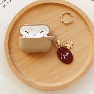 May include: A beige Airpods case with a gold chain and a burgundy leather keychain with the name "Kate" written in white.