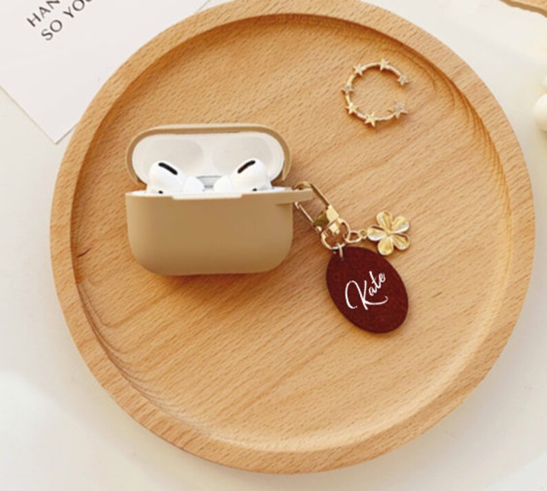 May include: A beige Airpods case with a gold chain and a burgundy leather keychain with the name "Kate" written in white.